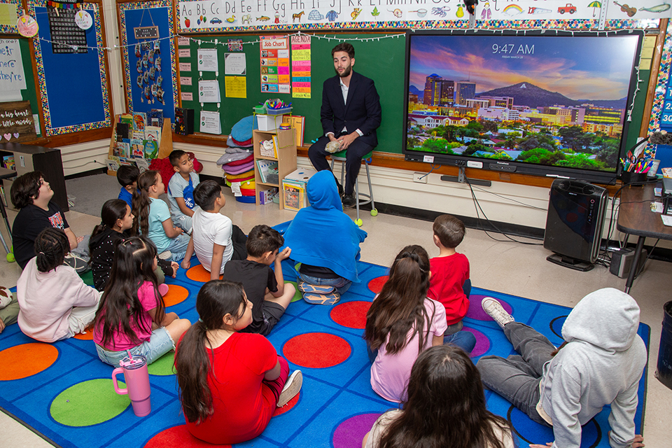 A man in a suit addresses a classroom of students sitting on the floor