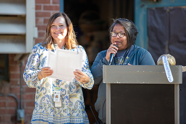 A woman smiles next to another woman standing behind a podium speaking 